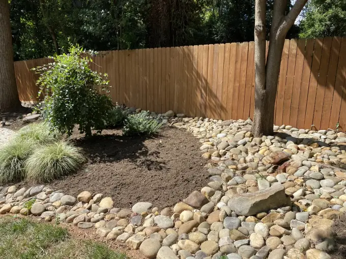 River-rock bed with ornamental grasses and shrubs along a wooden privacy fence