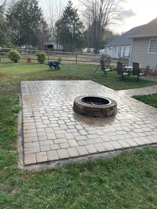 Square paver patio with a round stone fire pit set in a spacious backyard