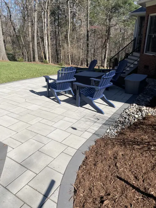 Paver patio with Adirondack chairs around a fire pit, edged with a river-rock border in a wooded backyard