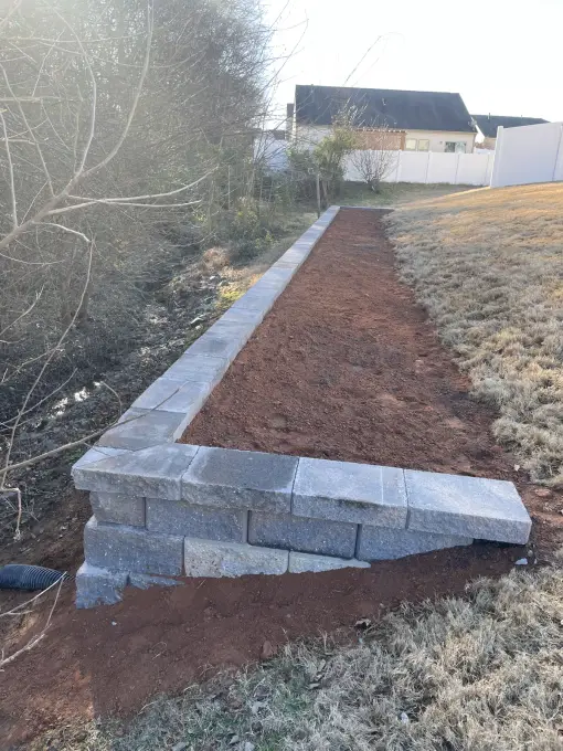 Stacked stone retaining wall built into a sloped yard with a fresh soil bed and drainage channel