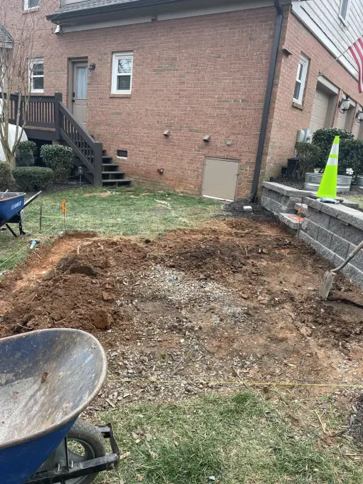 Retaining wall under construction with excavated soil and a wheelbarrow behind a brick home