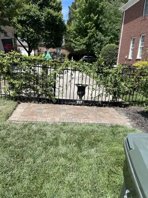 Brick paver landing pad set into the lawn at a black wrought-iron garden gate, bordered by fresh mulch