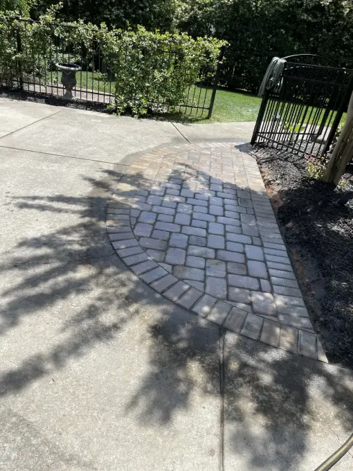 Circular cobblestone paver medallion inlaid into a concrete driveway apron beside a landscaped iron fence