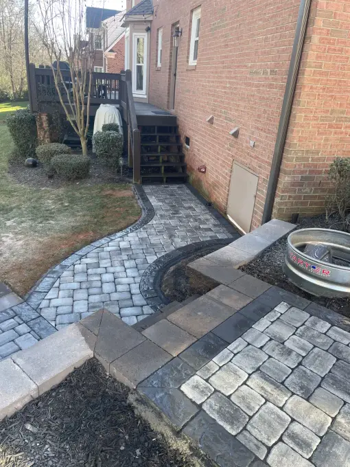 Cobblestone paver walkway with steps curving along a brick home toward a fire pit